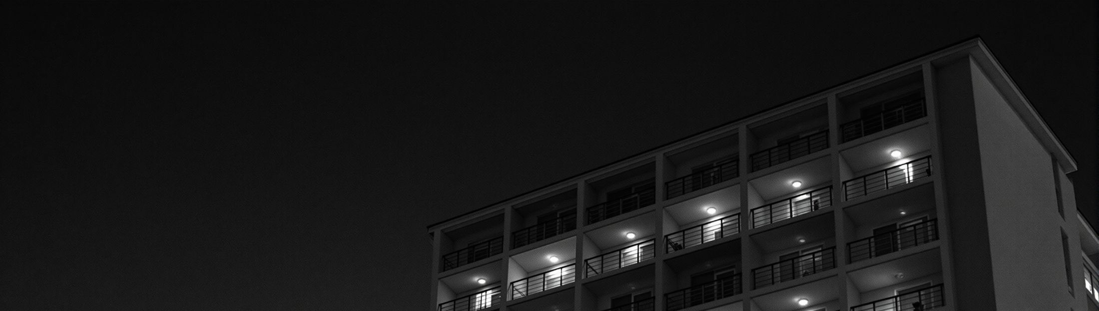 Fachada de un hotel modernista de los setenta al anochecer, con la luz cálida
        saliendo de las habitaciones por los balcones.